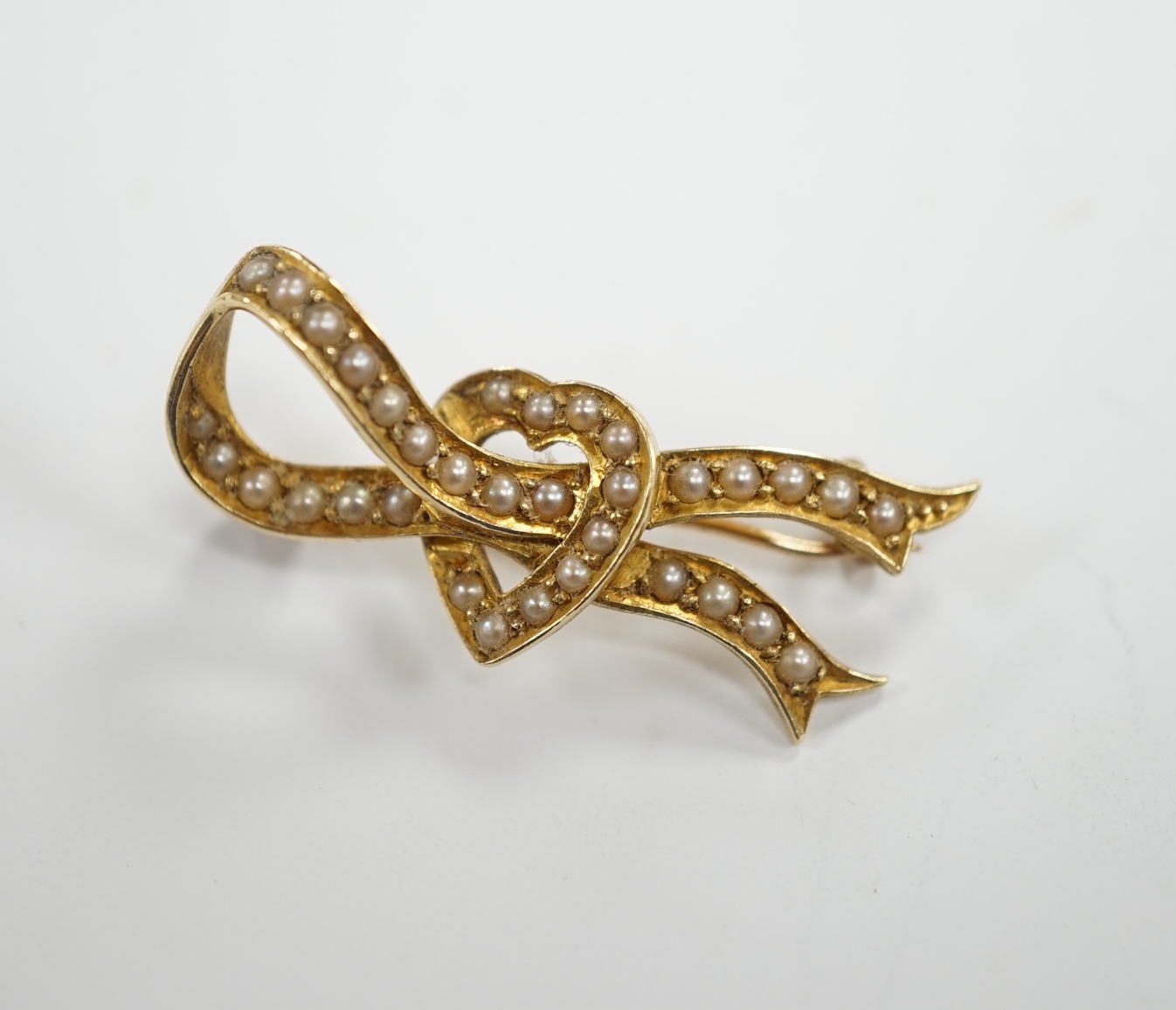 An Edwardian yellow metal and seed pearl cluster set ribbon and heart brooch, 34mm, gross weight 4.3 grams.